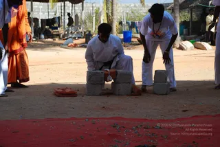 Outdoor photograph at a sandy ground under a thatched shelter with a red cloth spread in the foreground scattered with green leaves. A man in white kurta and white dhoti kneels between two stacked grey concrete blocks, with a terracotta brick and a red-ochre cloth placed beside him. A second man in a white shirt with a white dhoti and a beaded mala stands bent over, placing or adjusting a block. An orange-robed figure with a mala stands at the left edge, partly cropped. Plastic buckets, drying clothes and palm trees appear in the background.