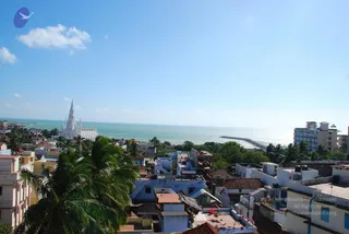 Elevated view over a coastal town toward the sea under a pale blue sky with scattered clouds. In the left middle distance a white church with tall spires rises above the rooftops, and palm trees fill the foreground. Flat-roofed houses in white, blue, cream and terracotta crowd the streets, with a breakwater jutting into the turquoise water on the right and a multi-storey white building beside it. No people, deities or ritual activity are visible.