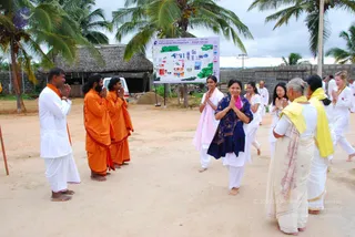 Outdoor photo at the Nithyananda Dhyanapeetam Bidadi Ashram on a dirt courtyard beside palm trees and a thatched shed. Three saffron-robed sadhus stand at left with hands joined in namaste, facing a line of devotees in white and cream clothes. A woman in the centre wears a dark blue shawl with a pink scarf and greets them with folded hands. Older women in cream sarees and yellow shawls stand at right, also with hands folded. Behind them a large signboard shows a map of the Bidadi ashram grounds. Another white-clad devotee stands near the far right. No identifiable a person is visible in the scene.