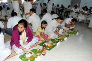 A hall with white tiled floor where many people sit on the floor and at low tables eating a communal meal from banana leaves. In the foreground a woman in a magenta cardigan and white-and-blue striped pants sits cross-legged, holding food with her right hand. Beside her a small girl in a yellow top and pink skirt eats from her leaf. Other seated participants wear white clothes and yellow lanyards, and a few people in white dhotis or saris stand at the back carrying steel vessels. Leaves hold rice, curries, pickles and vegetables, with a plastic bottle and steel tumblers placed among them. This appears to be an annadan style meal served during a retreat.