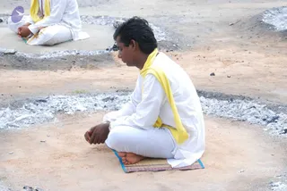 Outdoors on bare reddish-brown earth, a man sits cross-legged on a striped mat, eyes lowered, hands folded in his lap. He wears a white kurta and white trousers with a yellow shawl draped over his shoulders. Ash-grey ashes or burnt residue form a ring around the seated figure on the ground. Behind him, a second person in white with a yellow shawl sits on a low concrete platform, also in a meditative pose.
