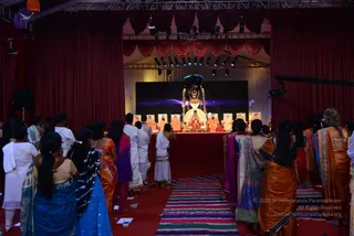 A photograph taken from behind a crowd of devotees facing a stage hung with red drapes and stage lights. Devotees in saris and white dhotis and kurtas stand on a striped runner rug. On a raised red dais, a central figure in orange robes sits before a large dark deity murti decorated with garlands, with several seated figures in saffron-orange robes on either side. A large screen glows purple behind the stage. The people in the foreground wear orange, blue, purple and white garments.