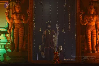 A photograph of a lit temple-style stage at night. Two large orange-toned guardian statues with multiple arms and crowns flank the central doorway. In the centre, a standing figure in a dark maroon garment with a gold necklace and a crown holds a silver trident staff, with a golden oil lamp base at the foot. Strings of small lights and hanging brass bells decorate the dark backdrop. The figure is a single person in a ceremonial pose; no identity is established by visible cues beyond costume and setting, so the figure is described by role only.
