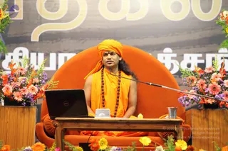 A man seated on a large orange-upholstered throne-style chair on a stage, wearing an orange turban, orange shawl and a long rudraksha mala. He sits behind a wooden table holding an open laptop and a small white device, with a glass of water beside him. A microphone stand extends toward him. Pink and orange gerbera daisies, orange lilies and purple flowers decorate both sides of the stage. Behind him is a large backdrop with Tamil script in white letters.