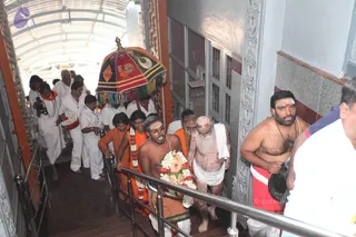 A procession ascends a covered stairway inside a building with grey stone-patterned walls and metal railings. A multicoloured canopy with tassels and rooster motifs is carried over the group. Leading figures wear orange and saffron cloths; one bare-chested man with a tilak and a wrapped white cloth carries a floral arrangement of roses, lilies and garlands on a tray. A shaved-headed man in a white dhoti walks bare-chested and bare-footed, his body smeared with white ash, and another man in an orange shawl wears a garland. White-clad attendants in white shirts and dhotis follow along the corridor. A bare-chested man on the right wears a rudraksha mala and red-bordered dhoti. The scene suggests a temple or ashram ritual procession.