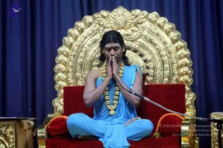 A seated man in a light blue dhoti-style garment and draped blue shawl sits cross-legged on a red cushioned throne before a large ornate gold arched backrest with floral and scroll carvings. He has long dark hair, a red bindi on his forehead, and a thick gold-beaded mala necklace. His palms are pressed together in anjali mudra with eyes closed. A microphone on a gooseneck stand stands at his side against a deep blue draped curtain, with a gold-trimmed table or stand at the left edge.