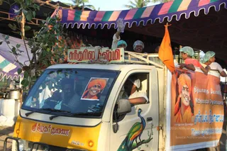 A yellow and white pickup truck with a peacock painted on its door carries volunteers for Thane cyclone relief. A man in a white shirt and hairnet waves from the truck bed, and other volunteers in white hairnets and caps stand along the side rails. A large banner on the truck side shows a portrait labelled "Paramahamsa Nithyananda" wearing an orange turban and smiling, with the text "Nithyananda Dyanapeetam" and "Thane Cyclone Relief". A smaller portrait in an orange turban is displayed on the windshield, and an orange flag flies from the truck. Colourful striped awnings and palm trees are in the background.