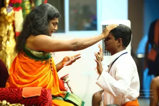 A photograph of a long-haired man in an orange and green silk shawl sitting on a red cushion, pressing his right palm to the forehead of a man in a white kurta with a blue cord and a dark sash. The seated man holds a folded white cloth in his left hand, with a rudraksha mala draped across his chest and a gold-toned ceremonial backdrop behind him. The recipient sits with palms folded in namaskar, eyes lowered, in what appears to be a blessing or initiation gesture. A small white air cooler and a seated figure in orange at the right edge are partly visible.