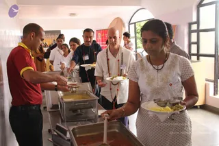 A buffet serving line in an indoor hall with pale walls and arched windows. A man in a red polo with yellow collar stands at left serving from steel chafing dishes of dal and curry. Behind him a man in a navy shirt with a red and beige lanyard holds a ladle. A bald man in a white shirt with long rudraksha malas and a lanyard holds a plate of rice and curry. In the foreground a woman in a white dress with gold embroidery and a lanyard carries a plate of rice, vegetables and dal. Several other attendees stand in line behind, and a banner reading Inner Awakening hangs in the background.