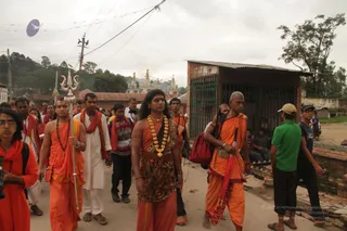 Outdoor street procession of saffron-clad sannyasis walking through a town lane near a Himalayan-style temple with tiered spires in the background. In the centre, a man with long dark swept-back hair wears a gold necklace of large beads, a leopard-print patterned top and an orange cloth draped over his shoulders, and walks behind a trident-topped staff. Beside him, a bald man in an orange robe holds a red umbrella and a red bag. Other followers wear orange and red shawls and white kurtas, and a woman in orange with glasses walks at the left edge. Onlookers stand on the right by a brick shed with a green metal gate, and power lines cross overhead.