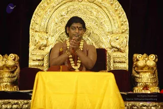 A seated man sits on an ornate gold throne with a carved arched back and two gold lion-shaped armrests. He wears an orange-red sash over bare shoulders, multiple gold necklaces and a dark rudraksha-style mala, with a red bindi on his forehead. He sits with palms pressed together in namaste behind a yellow draped table, with a microphone near his face. Two gold lion statues flank the foreground, and dark curtains form the backdrop. Swamiji (probable): long swept-back hair, saffron-orange garments, ceremonial gold necklaces and a rudraksha mala, seated on a throne, consistent with a satsang discourse.