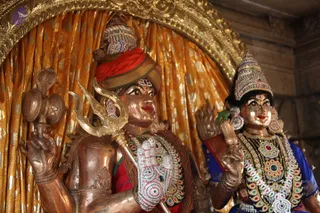 A photograph of two metal deity murtis (utsava images) in a temple shrine before a gold-patterned curtain with an ornate gilded arch. The figure on the left, a copper-toned deity murti, wears a red turban and red cloth, holds a golden trident (trishul) and a damaru-like drum with a snake motif, and has a bejeweled white-and-green ornamental armlet. The figure on the right, a female deity murti, wears a blue garment, a tall jeweled crown, a multi-strand gemstone necklace and a green leaf or flower in her hand. Both are richly adorned with jewelry and beaded ornaments. No physical people are visible.