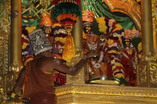 A priest in an orange-brown dhoti and sacred ash markings on his arm stands at a gilded shrine, reaching toward a row of decorated deity murtis. The murtis wear colourful flower garlands in orange, yellow, red and green, ornate silver crowns and gold jewellery. A golden kalasha-style pedestal and a tray sit in the foreground, with gold pillars framing the scene and green draped fabric and leaves in the background.