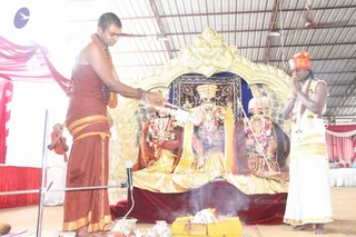 A priest in a maroon dhoti with a gold border and a sacred thread stands at left, pouring liquid from a white vessel over three deity murtis seated on a gold-draped platform. The central murti wears a tall gold crown and garlands; the murtis on either side wear garlands and ornate jewellery. In the foreground a small fire pit with a yellow box and offerings burns, sending up smoke, indicating an abhishekam with homa. At right, a man in an orange headcloth and white dhoti stands with palms pressed together in prayer. Behind them, a gold arch, red and pink draped fabric, and a microphone on a stand frame the shrine under a metal-roofed pavilion.