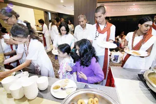A food serving line inside a hall with a pink banner reading Nithyananda Gallery. Devotees in white clothes with red-orange shawls and rudraksha malas serve and collect food from steel trays and buffet tables. A young shaved-headed man in white, wearing a red-orange shawl and beaded necklace, holds a white plate of food. A girl in a purple top and a girl in a pink patterned top queue for vada-style fried snacks and a small bowl of sauce. Stacks of white bowls sit on the table in front, and an older man in white watches from behind. No a person candidate is visible in this frame.