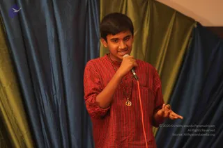 A young man stands before green and blue draped silk curtains, holding a microphone in his right hand and gesturing with his left as he speaks. He wears a red and white striped kurta with rolled sleeves, a wooden rudraksha mala, and a small round pendant on a cord. He is smiling and appears to be addressing an audience.