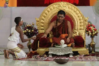 A photograph of a seated man on an ornate gilded throne with a red cushioned seat, wearing a saffron-orange robe, a long rudraksha mala hanging to his chest, and a red bindi on his forehead. He extends his hand over a silver pedestal holding a silver bowl and green leaves. In front of him kneels a young boy with a shaved head and a white horizontal mark, wearing white dhoti with gold borders, holding out an offering. Red and purple flowers in brass vases frame the throne, and a copper vessel sits on the floor.