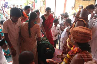 Crowded outdoor temple-area scene at a ceremony. In the foreground a woman in a green sari bows low, head bent over a mat laid on the ground, while a woman in a peach-coloured sari with a long braid steadies her. Behind them a woman in a red sari and gold necklace watches closely. At right, a bald man with a brown-and-orange turban, wearing orange-saffron cloth and rudraksha malas, stands among other devotees in white and orange garments; a garland of orange and red flowers hangs around his neck. Onlookers, including a young boy in a light shirt and a man in white dhoti, fill the background under a tent-like canopy.