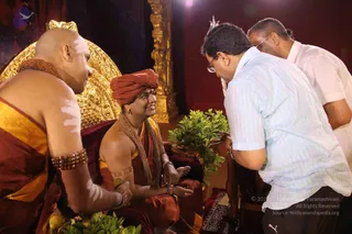 A photograph of a satsang setting with a golden ornate throne against a red backdrop. Seated on the throne is a man in an orange-red turban, orange-red and gold robes, with layered beaded malas across his chest and beaded bracelets on his wrists, speaking with a smile. Beside him on the left sits a shaved-headed man in an orange-and-gold shawl with sandalwood markings on his arms and chest and a rudraksha mala. In the foreground a devotee in a light-blue striped shirt and glasses bends forward, offering hands toward the seated figure, while another man in a white shirt stands behind. Green leafy foliage sits at the base of the throne.