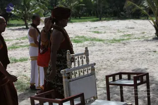 Outdoor photo on a sandy clearing with palm trees and patchy grass behind. In the centre a woman in a dark red turban and a dark patterned skirt, with gold necklaces and bangles, stands beside an ornate white and gold carved chair with a white cushion and tassels, her arm raised. To the left, a man in a white dhoti with gold borders and a shaved-headed monk in saffron-red robes stand near a red carrying bag. Two wooden stools sit in the foreground.