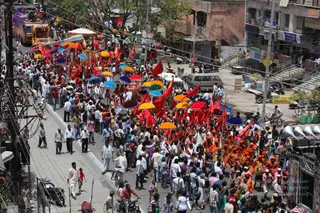 Aerial view of a dense street procession in an Indian town, with a large crowd moving down the road between shopfronts and parked motorbikes. Many people carry red flags and open umbrellas in yellow, orange, red, blue and green. Several figures in saffron and orange robes walk near the front, while others wear white kurtas and dhotis. A decorated vehicle with a golden canopy is visible at the top left, and loudspeakers are mounted on a stand at the right edge. Ceremonial procession with red flags and umbrellas on a city street.