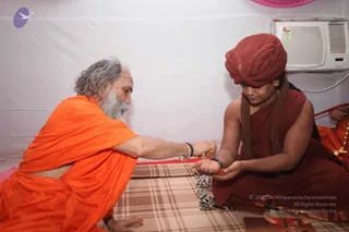 Indoors against a white wall with a window air conditioner, two men sit on a checked cloth-covered mat. On the left, an older man with grey curly hair and a long grey beard wears an orange saffron shawl and bangles, and leans forward while holding a small object toward the younger man. On the right, a younger man wears a maroon-red turban, a maroon robe and a rudraksha-style beaded mala, and holds out both cupped hands to receive it. Another person in orange sits partly visible at the right edge. A small pink object hangs at the top corner.
