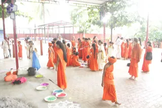 A photograph of an open-sided pavilion with a tiled floor and red-painted pillars, where many people in saffron-orange robes gather. Several women and young girls in orange saris and some with black hair in long braids stand and move about, while a few figures in white clothing are also visible. One young figure in the foreground wears an orange turban and orange robes. Small shallow bowls on the floor hold coloured powders in green, pink, purple and blue, and an orange cloth bag and a black bag sit near the left edge. Trees and a red boundary wall are visible beyond the pavilion.