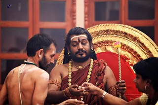 A photograph of a ritual in progress before a gilded, carved golden backdrop with a red cushion. A bare-chested man in white sacred marks on his shoulders and a sacred thread stands at left with his back to the camera. At centre, a man with long swept-back hair, white forehead band and red mark, wears a rust-red wrap, a heavy gold bead necklace, rudraksha-style beads on his arms, and earrings; he looks down and holds a small object in his cupped hands. At right, a woman in a dark top and silver bracelet extends her hand toward him, and a flower-decorated staff rises beside the throne. Wooden latticed windows form the background. Multiple elements suggest a puja or darshan setting.