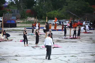 A photograph of an outdoor paved courtyard where a mallakhamba-style pole yoga demonstration is underway. Several tall wooden poles stand upright, with practitioners performing postures on and beside them. A woman in black athletic clothing climbs a pole while others in black stand on mats and balance on pole-side mats. Groups of people in saffron-orange robes stand in the background near parked cars. In the foreground, a man in a white shirt and dark trousers with a red cloth draped over his shoulder walks across the courtyard. Trees, a stone wall and a small signboard frame the scene.