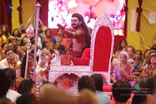 A photo of a long-haired man in an orange-brown shawl and rust-red cloth, seated on an ornate silver-and-red throne with swan carvings, one arm raised in a gesture toward a large crowd. A silver staff with a lotus-topped finial stands at the left, and a silver pitcher sits near the throne. Behind him a large screen shows a bright, colourful image, with red and gold draped fabric framing the hall. Many seated devotees fill the foreground and background, several women in saffron, orange and purple saris with garlands and beaded necklaces, and men in white and orange. a person: the long dreadlocked hair, saffron shawl and ceremonial throne match the presentation of a teacher on stage, but the face and posture are not clearly resolved, so the identity is uncertain.