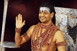 A photograph of a man with a white forehead band and a dark vertical tilak, long dark hair swept back, and small gold earrings, raising one hand in blessing. He wears layered rudraksha-style beaded necklaces in reddish-brown with gold and jewelled pendants, matching beaded armlets, and is bare-chested. Behind him is an ornate silver-coloured throne with embossed floral and heart motifs, and a silver pole stands at left. Swamiji (probable): the qualifying cues are the swept-back long hair, layered rudraksha malas, ornate beaded adornments and the blessing gesture before the ornate silver throne.