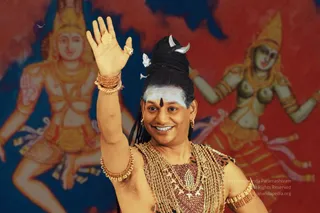 A smiling man with a long black topknot adorned with silver crescent and butterfly ornaments, white vibhuti stripes and a black bindi on his forehead, raises his right hand in a blessing wave. He wears heavy gold necklaces, gold bangles and armlets, and a maroon-and-gold draped garment. Behind him, painted mural figures of a deity in gold and red and a dancing figure in red and blue fill the background. a person: the figure has long hair and the cues of gold ornaments and sacred ash, so this is a probable depiction of the a person presence in the scene.