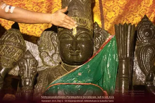 A photograph of a dark stone deity murti draped in a green cloth with a red border, set against a yellow floral backdrop. A bare hand with white and blue marks on the forearm touches the forehead of the murti, where a yellow mark is applied. Carved stone elements, including a tall ornate crown and sculpted side pieces, frame the figure. The caption identifies the setting as Nithyanandeshwara Paramashiva Devalayam, Adi KAILASA, during Navaratri 2021, Day 6, Sri Rajarajeshwari Abhishekam and Kalasha Yatra.