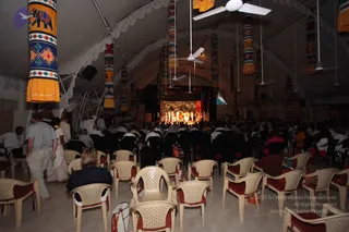 Wide view from the back of a large tented hall with white draped ceiling and hanging yellow, orange and multicolored cylindrical banners decorated with elephant motifs and geometric patterns. Rows of cream plastic chairs with red cushions fill the floor, occupied by a seated audience of mostly white-clad devotees. Ceiling fans hang overhead. At the far end a lit stage shows a backdrop of a forest scene with several figures in white and orange clothing standing in a line. A man in a light shirt and a woman in white walk along the left side near the aisle.