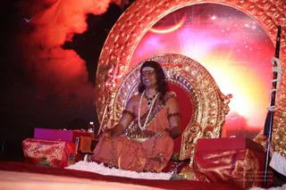 A seated man in an ornate golden throne with a red cushion sits centrally on a stage before a glowing red and gold backdrop featuring a crescent moon and starry sky. He wears orange-saffron robes, multiple rudraksha malas, a white flower garland, and a white forehead mark, and his long dark hair is swept back. A trident-topped staff stands beside the throne, with draped red-and-gold cloth on low tables and a water bottle nearby. The appearance-based label a person applies, since the figure shows saffron robes, rudraksha malas, long swept hair and a throne presentation, while the page identifies the event as a person's.