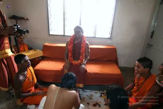 A photograph inside a small plain room with white walls and a barred window. A man in a red-orange robe with a bright orange and yellow flower garland sits on an orange-covered couch, smiling, with a bindi on his forehead and gold armlets. Before him sits a shaved-headed priest in white, bare-backed with a sacred thread, in front of a white cloth with offerings. Around them, several young people in orange robes and rudraksha malas sit on the floor, one holding a tray. A standing man at left in reddish-orange clothes holds a camera, and a small wooden shelf with a bag is on the wall.