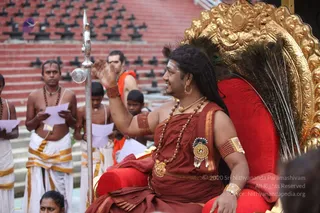 A man seated on an ornate golden throne with a red cushioned seat and a peacock-feather backrest raises his right hand in blessing. He wears a maroon-red robe with a gold border, layered rudraksha malas, gold necklaces, a gold brooch, gold armlets and red bangles, and has long dark hair with a tilak on his forehead. Beside him stands a tall ceremonial staff topped with a metal finial. Around the throne, several men and boys in white dhotis with gold borders, some bare-chested and wearing rudraksha malas, hold sheets of paper, and a man in orange cloth stands behind. Red-tiered stone steps form the background. a person is seated on the throne, identified by his long swept hair, ornate gold jewellery and rudraksha malas.