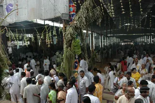 A large crowd gathers under a long corrugated-roof pavilion decorated with hanging green palm-leaf festoons and a tall palm-frond and banana-bunch arch in the centre. Most people wear white garments: men in white shirts and dhotis, women in white saris and some in yellow or other coloured saris. Many stand close together in the aisles and at the edges, with a few older women and men seated or standing near the front. A striped cloth panel and a red and blue banner hang from the central pole. Hundreds of people fill the open hall, with more figures receding toward the far end under rows of hanging lights.
