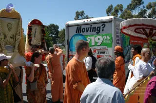 A street procession on a sunny day beside a white U-Haul truck with a 'Moving?' advertisement. Several people in orange robes walk alongside, including a man in orange with a shaved head and a woman in an orange head covering. Two devotees carry framed portrait images of spiritual figures decorated with white flower garlands, one with orange marigold garlands. A white-and-pink umbrella canopy appears at right, and a man in white kurta holds a pole. Trees and blue sky are in the background.