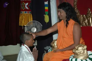A photograph of a seated man in saffron-orange robes, with long dark swept-back hair and a small tilak on his forehead, seated on an ornate gold and red throne. He rests one hand on the head of a kneeling young boy in white clothes with a red-and-white cord, whose hands are pressed together in anjali. Behind them hang a red and gold embroidered cloth, a dark curtain, a white fan and a green, white and yellow garland. Gold lion-like armrests and white-and-green flower garlands decorate the throne. The scene suggests a blessing given during a gathering.