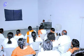 A photograph inside a plain whitewashed hall with a blue-grey wall and a dark cloth hanging over a window. Rows of white plastic chairs face a small table at the front holding a black amplifier speaker and a laptop. A man in a white shirt stands behind the table speaking. Around a dozen seated attendees, mostly seen from behind, wear white clothes with orange shawls or wraps, and several have long black hair in braids. Two standing electric fans sit on the right side of the room.