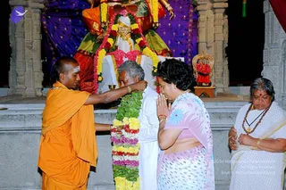A temple scene at night with a deity murti draped in purple and gold fabric and garlanded with flowers. In the foreground, a man in orange robes with a white tilak on his forehead stands at left, placing his hand on a large flower garland. Beside him, an older man in white clothes bows his head near the garland. A woman in a pink and white polka-dot sari stands with palms pressed together in namaste, and an older woman in a white sari with a rudraksha mala looks on at right. A small ornate gold throne with red flowers sits to the right. Stone pillars frame the shrine.