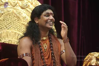 A smiling man with long, swept-back dark hair sits on a red velvet seat in front of an ornate gold throne-back. He wears an orange-saffron cloth draped over one shoulder, a red bindi on his forehead, a gold pendant necklace, a gold torque, and long multi-strand rudraksha malas across his chest. Beaded bracelets and armlets adorn his arms, and he gestures with one raised hand while speaking. A deep magenta curtain forms the background. a person is shown in this photo, seated in conversation during a satsang setting.