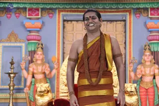 A smiling man stands centrally on a golden-throne stage before a painted temple backdrop with carved pillars. He wears a rust-orange and gold-bordered dhoti-style wrap, a matching shoulder cloth, gold-banded armlets and a long multi-strand rudraksha-style mala with a red bindi on his forehead. A small headset microphone is attached to his face. Behind him stand two large painted deity statues with raised arms and crowns, flanked by golden lion-shaped seats. A brass oil lamp stands at the left edge. a person appears in person, identified by the saffron-orange clothing, rudraksha mala and ornate gold armlets, with a microphone for a satsang-style discourse.