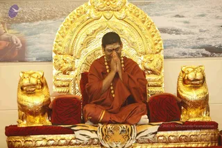 A seated man in saffron-orange robes sits cross-legged on an ornate golden throne with a high arched backrest and carved lion heads at each side. He has his palms pressed together in namaste at his chest, and wears a rudraksha mala with gold-tone beads and a vibhuti or tilak mark on his forehead. A tiger-striped cloth covers the seat, and red velvet cushions line the armrests. Behind him a framed photograph shows a sea and clouds.