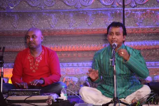 A photograph of two men seated cross-legged on a stage in front of an ornately carved stone-patterned backdrop lit in red and blue. The man on the left is bald, wearing a red kurta with embroidered neckline and a dark wristwatch, and sits behind a keyboard instrument with his hands on it. The man on the right has short dark hair and wears a green striped kurta with white dhoti-style trousers; he sings or speaks into a microphone on a stand while gesturing with his right hand. Water bottles and cables lie on the stage floor. Nothing here shows a throne, crown, saffron clothing or rudraksha, so no a person candidate is established by appearance.
