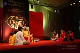 A stage event photographed from the front. A woman in a yellow and gold sari sits on an ornate silver-and-gold chair draped in red, flanked by a small table with a silver bowl. Before her on the red stage, a young man in a white kurta sits cross-legged with two boys in yellow and orange robes, while three women in red and white sit on the right, one holding a phone. Behind them a large banner reads Nithyananda Yogic Sciences Presents Manifesting Shaktis, showing a bearded man in a red turban with a garland, raising his hand inside a gold lotus-and-triangle emblem. Red drapes and stage lights frame the scene.