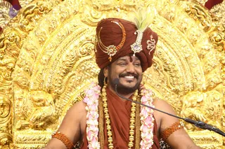 A smiling man sits in front of an ornate golden throne backdrop with embossed floral and scrollwork patterns. He wears a rust-red turban decorated with a crescent-shaped gold ornament, a jewelled brooch and a green feather, plus a red-orange draped cloth, layered gold chains and pink-and-white flower garlands. Orange rudraksha bead bracelets are on both upper arms, and a microphone stands in front of him. Swamiji (probable): long-haired figure with saffron-red turban and ceremonial gold necklaces, seated speaking before the throne.
