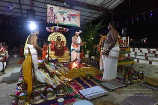 Night ritual at an open-sided temple pavilion with a corrugated roof and string lights. A seated figure in red-orange robes and a rudraksha mala sits on an ornate gold throne with a red cushion beneath a striped canopy. Before him, a fire burns in a raised golden fire pit as a bare-chested priest in white dhoti and pink head-cloth stands with a ritual vessel. Two bare-chested attendants in white dhotis and yellow shawls stand at the sides, one with garlands, and another stands near a tray of fruit and flowers. Brass vessels, marigold and rose garlands, a kalasha-style arrangement and a deity poster on the back wall complete the scene, which appears to be a homa or yagna. Above the poster, a world map and printed text are visible. Several figures in orange garments sit on the ground at the left and right edges.