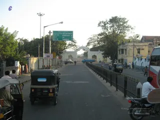A street scene in a city, viewed from a roadway with a low metal railing on the right. A green road sign reads Mysore Palace, Zoo, Chamundi Hill with Kannada text above. A white decorative gateway arch stands in the distance beneath trees. Several auto-rickshaws in black and blue are visible in the foreground, with motorcycles and pedestrians on both sides of the road and a red bus on the right edge. No temple ritual, discourse or ashram activity is shown, and no person matching a person candidate is visible.
